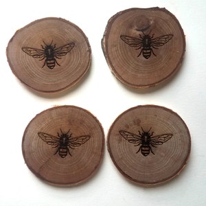 May include: Set of four wooden coasters with a bee design. Each coaster is round and has a natural wood finish. The bees are black and are centered on each coaster.