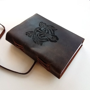 May include: A dark brown leather journal with a debossed tiger head design on the cover. The journal is bound with visible stitching and has a leather strap closure. The pages are white.