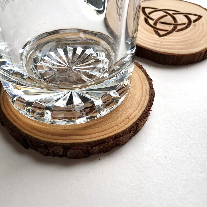 Puede incluir: Primer plano de un vaso transparente con un dise&ntilde;o de vidrio cortado que descansa sobre un posavasos de madera. El posavasos est&aacute; hecho de una rodaja de madera natural con un borde de corteza. El posavasos tiene un dise&ntilde;o de nudo celta tallado en la superficie.