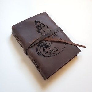 Op de afbeelding: Een bruin leren notitieboek met een vuurtoren- en golfontwerp. Het notitieboek is gesloten met een leren band. Het vuurtoren- en golfontwerp is zwart. Een leuk cadeau voor schrijvers en liefhebbers van de zee.