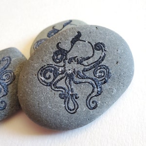 Può includere: Una pietra grigia con un disegno a inchiostro nero di un polpo con tentacoli arricciati.