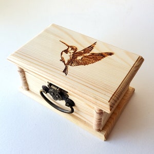 Puede incluir: Una pequeña caja de madera con tapa con bisagras y asa de metal. La parte superior de la caja está decorada con una imagen de colibrí quemada en madera.