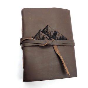 May include: A brown leather journal with a mountain range design embossed on the front cover. The journal has a leather strap closure.