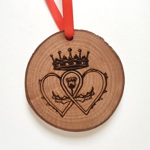 May include: A wooden ornament with a crown and two intertwined hearts with a thistle in the center. The ornament is brown and has a red ribbon attached.
