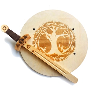 May include: A wooden toy sword and shield set. The shield is round and features a Celtic knot design with a tree of life in the center. The sword is made of wood and has a brown leather handle.