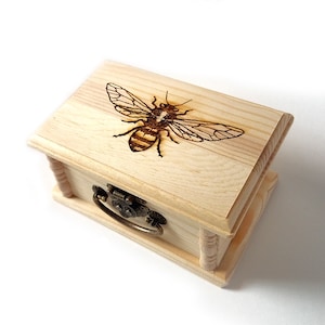 May include: A small wooden box with a hinged lid and a brass latch. The top of the box is decorated with a woodburned image of a bee.