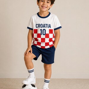 May include: A white t-shirt with a red and white chequered pattern, the text CROATIA, and the number 10 in dark blue, paired with dark blue shorts, white socks with two dark blue stripes, and white trainers, standing with a black and white football.