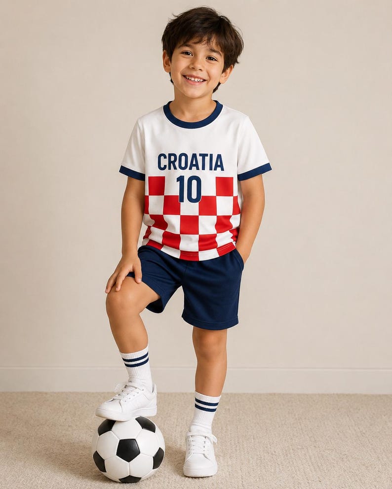May include: A white t-shirt with a red and white chequered pattern, the text CROATIA, and the number 10 in dark blue, paired with dark blue shorts, white socks with two dark blue stripes, and white trainers, standing with a black and white football.