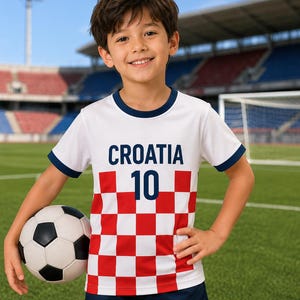 May include: A white short-sleeved football shirt with a red and white chequered pattern on the lower half, featuring the text CROATIA and the number 10 in dark blue, paired with dark blue shorts and a black and white football.