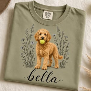 Può includere: Una t-shirt girocollo color verde salvia con la grafica di un cane dorato che tiene in bocca una pallina da tennis gialla, circondato da fiori di campo disegnati a tratto. Il nome "bella" è stampato in corsivo nero sotto l'illustrazione.