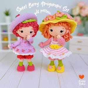 Peut inclure: Deux poupées amigurumi crochetées, l'une dans une robe et un béret violets, l'autre dans une robe et un chapeau de soleil roses. Les poupées ont des cheveux bouclés et sont étiquetées "Sweet Berry Amigurumi Set pdf pattern". Les poupées sont sur une surface blanche.