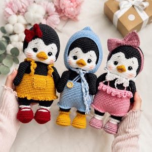 Può includere: Tre bambole pinguino all'uncinetto con corpi neri e bianchi e becchi arancioni. La bambola a sinistra indossa un vestito giallo e scarpe rosse. Quella centrale indossa un completo blu con cappuccio e stivali gialli. Quella a destra indossa un completo e un fiocco rosa.