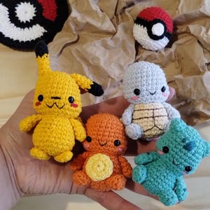 May include: Four small crocheted figures including a yellow Pikachu, an orange Charmander, a light blue Squirtle, and a teal Bulbasaur, accompanied by a crocheted Poké Ball and a flat circular Poké Ball design in the background.