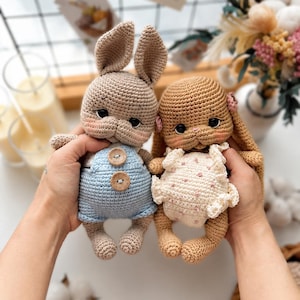 May include: Two handmade crochet stuffed animals shaped like rabbits. The left rabbit is light brown with blue overalls featuring two wooden buttons. The right rabbit is tan with a cream-colored ruffled romper featuring small pink dots and pink ear accents.