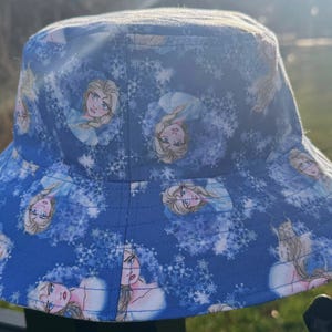 Może przedstawiać: Niebieski kapelusz typu bucket z powtarzającym się wzorem postaci z kreskówki z blond włosami i niebieską sukienką, otoczonej białymi płatkami śniegu. Kapelusz jest wykonany z tkaniny i ma szerokie rondo.