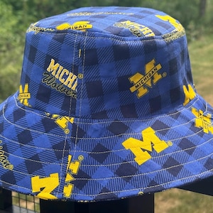 May include: A blue and dark blue plaid bucket hat featuring the yellow Michigan Wolverines logo and the text Michigan Wolverines printed repeatedly across the fabric. The hat has a flat top, a downward sloping brim, and visible yellow stitching.