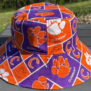 May include: A bucket hat featuring a repeating grid pattern of orange and purple squares. The design includes the Clemson tiger paw logo and the text CLEMSON printed in white. The hat has a wide, downward-sloping brim and a flat crown.