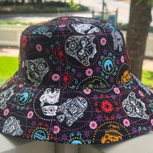 May include: A black bucket hat featuring a colorful repeating pattern of white, yellow, and blue Star Wars character illustrations, including Darth Vader, R2-D2, and Stormtrooper helmets, interspersed with pink flowers and red circular symbols.