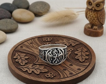 May include: A silver ring with a detailed dragon design. The ring features a central dragon motif with outstretched wings, surrounded by intricate patterns. The ring is displayed on a wooden coaster with carved leaf and floral designs.