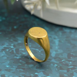 May include: A gold signet ring with a smooth, oval face. The ring has a polished finish and a classic design. The band tapers towards the back. The ring is shown on a blue-green surface.