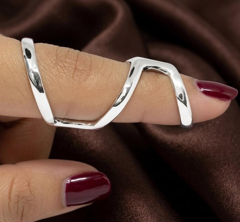 Puede incluir: Un anillo plateado, con un dise&ntilde;o geom&eacute;trico en forma de V, rodea dos dedos. El anillo se lleva en una mano con esmalte de u&ntilde;as burdeos. El fondo es una tela marr&oacute;n oscuro.