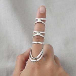 May include: A silver-colored finger ring with multiple connected bands. The ring has a unique design, with a curved lower section and several horizontal bars. The ring is worn on a finger, showcasing its intricate details and modern aesthetic.