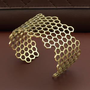 Könnte beinhalten: Ein goldfarbenes Armreif-Armband mit Wabenmuster. Das offene Armband weist eine Reihe von miteinander verbundenen sechseckigen Formen auf. Das Armband ist aus Metall gefertigt und hat eine glänzende Oberfläche. Der Schmuck wird auf einer braunen Oberfläche präsentiert.