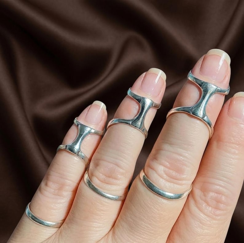 Puede incluir: Anillos de dedo plateados en varios dise&ntilde;os. Los anillos se llevan en los dedos y tienen un aspecto &uacute;nico y moderno. Los anillos est&aacute;n hechos de un material met&aacute;lico brillante y se muestran sobre un fondo marr&oacute;n.