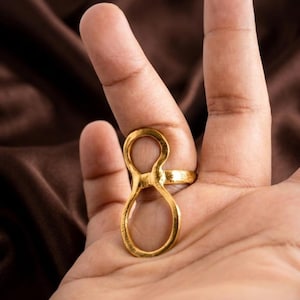 Op de afbeelding: Een goudkleurige metalen ring met een dubbel lusontwerp dat verticaal langs de vinger loopt. De band is verbonden met twee gestapelde, open cirkelvormen, waardoor een langwerpig figuur-acht silhouet ontstaat met een gladde afwerking.