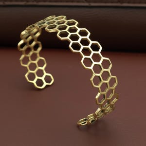 Könnte beinhalten: Goldfarbenes Armreif-Armband mit Wabenmuster. Das offene Armband hat eine Reihe von miteinander verbundenen sechseckigen Formen. Der Schmuck besteht aus einem glänzenden, metallischen Material und wird auf einer braunen Oberfläche präsentiert.