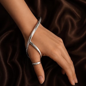 May include: A polished silver-tone hand bracelet featuring a smooth, curved metal frame that extends from the wrist across the back of the hand, connecting to a simple, solid band that encircles the thumb for a secure and continuous fit.