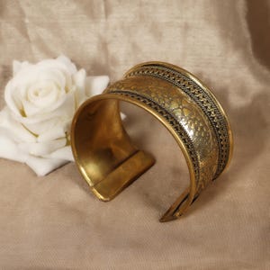May include: A wide, open brass cuff bracelet with intricate floral and geometric patterns. The cuff has a slightly curved shape and a polished finish. A white rose is in the background.