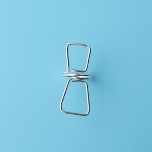 Könnte beinhalten: Eine einzelne, glänzende, silberfarbene Metall-Wäscheklammer vor einem einfarbigen hellblauen Hintergrund. Die Wäscheklammer hat ein einfaches, geometrisches Design mit zwei quadratischen Enden, die durch eine Spiralfeder verbunden sind.