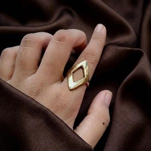 Pode incluir: Um anel de tom dourado com uma peça central geométrica aberta em forma de diamante. A banda é lisa e polida, exibindo uma superfície metálica reflexiva que se estreita ligeiramente onde se conecta à estrutura central em forma de losango.