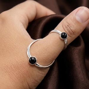 May include: A silver-toned metal ring featuring two circular black stones. The design consists of a larger band that wraps around the base of the thumb and a smaller band that sits near the knuckle, connected by a thin silver metal bridge.