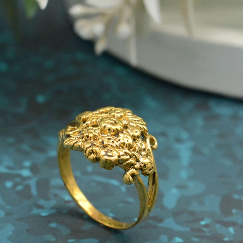 K&ouml;nnte beinhalten: Ein goldfarbener Ring mit floralem Design. Der Ring hat einen detaillierten, strukturierten Blumencluster an der Oberseite mit einem zarten Band. Der Ring ist vor einem teal-farbenen Hintergrund platziert.
