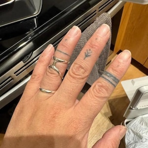 Puede incluir: Una mano con varios anillos de plata y tatuajes en los dedos. Un anillo es una banda simple, y otro tiene un dise&ntilde;o abierto &uacute;nico. Los tatuajes incluyen una l&iacute;nea fina, un peque&ntilde;o dise&ntilde;o floral y una serie de l&iacute;neas alrededor de los dedos.