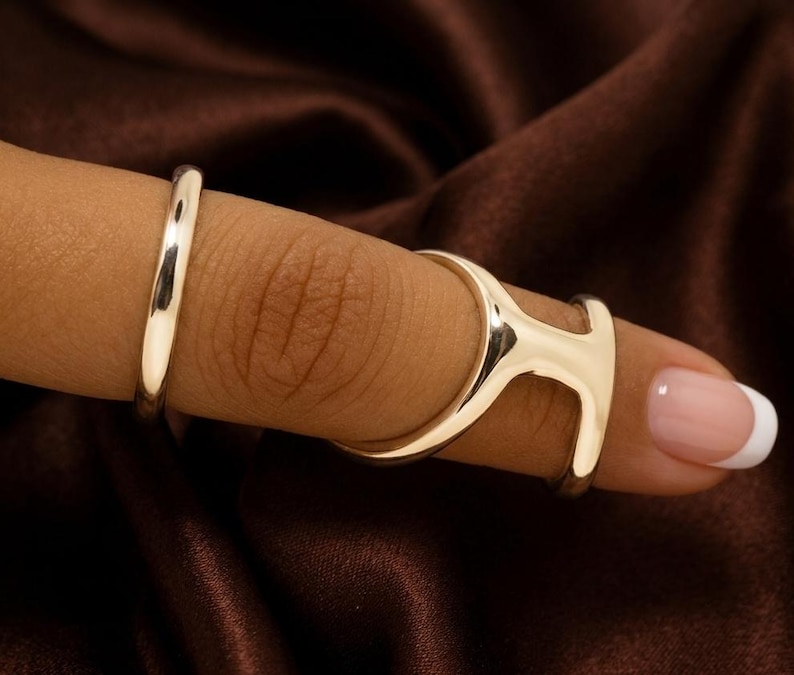 Puede incluir: Primer plano de tres anillos plateados en un dedo. Uno es una banda simple, y los otros dos est&aacute;n conectados, formando un dise&ntilde;o geom&eacute;trico &uacute;nico. El fondo es una tela marr&oacute;n oscuro.