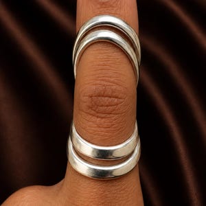 May include: Silver finger rings. The rings are smooth and curved, with a polished finish. The rings are worn on a finger, and the image is set against a brown background.