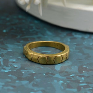 Könnte beinhalten: Ein goldfarbener Ring mit einem geometrischen Muster aus verbundenen Pfeilspitzen um das Band. Der Ring besteht aus massivem Metall und liegt auf einer blauen Oberfläche.