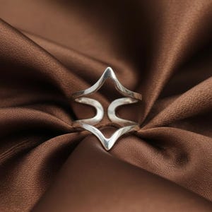 May include: A silver ring with a geometric design. The ring features a central diamond shape with curved accents. The ring is displayed on a brown satin fabric.