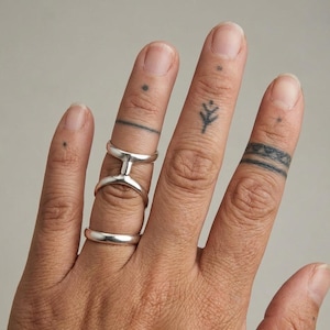 Puede incluir: Una mano que muestra tres anillos de plata. Uno es una banda simple, otro tiene un dise&ntilde;o de doble banda distintivo, y el tercero es un anillo de conexi&oacute;n. Los dedos tambi&eacute;n tienen peque&ntilde;os tatuajes.