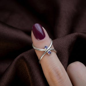 Könnte beinhalten: Ein silberner Ring mit einem X-förmigen Design, der zwei kleine, runde, violette Edelsteine ​​aufweist. Der Ring wird an einem Finger mit burgunderfarbenem Nagellack getragen. Der Hintergrund ist ein dunkelbrauner Stoff.