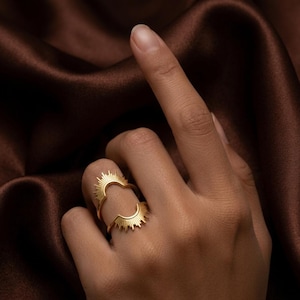Könnte beinhalten: Ein goldfarbener Ring mit Sonnenstrahlen-Design. Der Ring ist in zwei Teile geteilt, einen für die Oberseite und einen für die Unterseite des Fingers. Der Ring wird auf einer Hand vor braunem Hintergrund getragen.