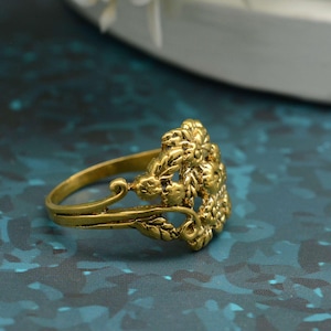 K&ouml;nnte beinhalten: Ein goldfarbener Ring mit floralem Design. Der Ring hat ein detailliertes, erhabenes Muster aus Blumen und Bl&auml;ttern. Das Band ist ein einfaches, glattes Gold. Der Ring ist vor einem dunkelblauen Hintergrund platziert.