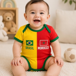 Może przedstawiać: Uśmiechnięte dziecko w żółto-czerwonym body z zielonymi wykończeniami. Body przedstawia flagi Brazylii i Maroka z napisami "BRASIL" i "MOROCCO" poniżej. Dziecko siedzi na białym dywanie.