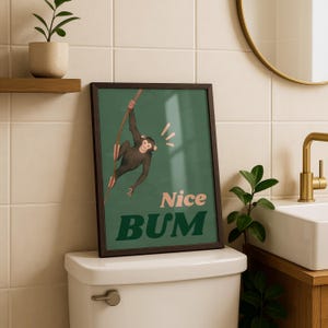 May include: Framed wall art featuring a cartoon monkey hanging from a branch against a green background. The text "Nice BUM" is displayed in pink and green. The artwork is in a dark brown frame.