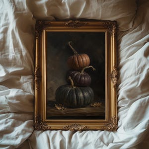 May include: A rectangular painting in an ornate gold frame featuring a vertical stack of three pumpkins in shades of orange, deep purple, and dark green. The pumpkins rest on a wooden surface with small dried leaves scattered at the base.