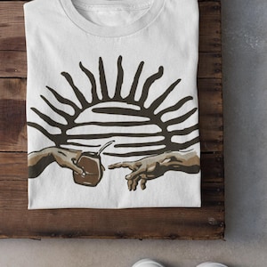 Può includere: T-shirt bianca con una grafica marrone raffigurante due mani che si tendono l'una verso l'altra, una che tiene una tazza marrone con una cannuccia e un disegno a forma di sole sopra. La maglietta è piegata su una superficie di legno.