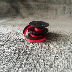 May include: A stack of three black and red typewriter ribbon spools. The spools are stacked on top of each other, with the red ribbon visible between the black spools. The ribbon is partially unraveled.